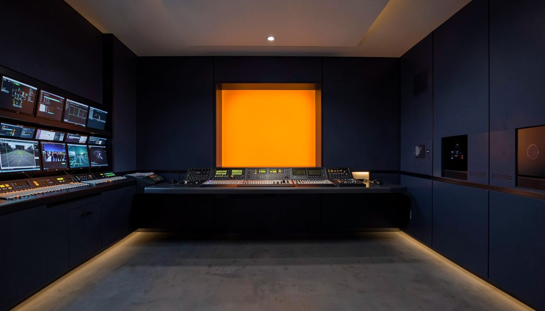 A clean architectural photograph of a broadcast studio control room with strong symmetry, deep navy and charcoal tones, warm amber accent lighting, and a wall of monitors glowing softly.