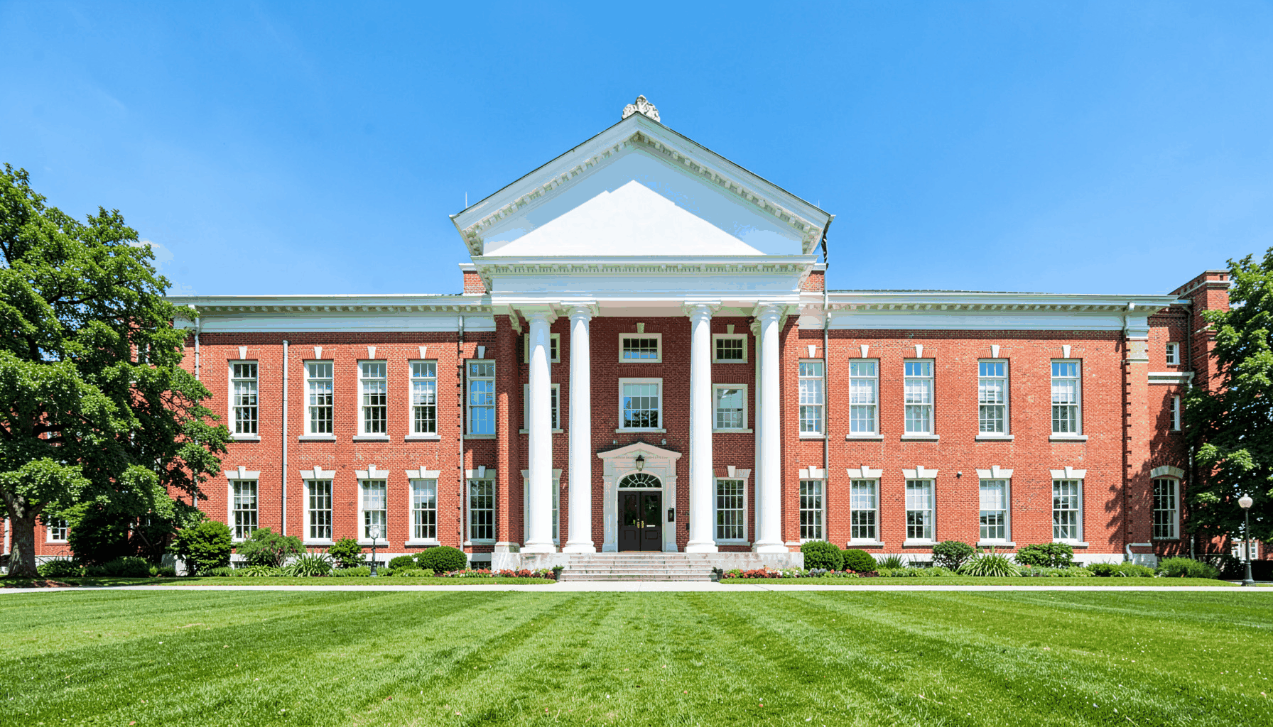 A wide-angle view of a stately red-brick academic building with white columns and a manicured green lawn under a bright blue sky, conveying tradition and campus serenity.