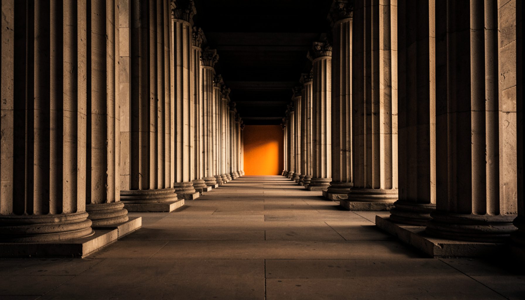 A grand colonnade of tall stone pillars receding in symmetrical perspective, warm sepia light washing across fluted columns, deep charcoal shadows pooling between them, conveying timeless strength and quiet authority.