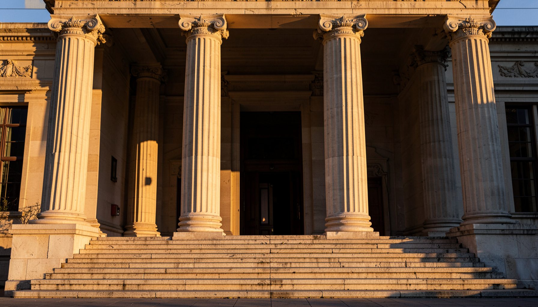 A stately neoclassical building facade with tall columns and broad stone steps, bathed in warm afternoon light, deep shadows accenting the symmetrical architecture.